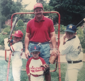 A man stands between two boys holding baseball bats, and a younger boy holding a baseball glove. All are wearing baseball caps and uniforms.