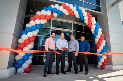 Four men in business attire stand in front of a building entrance, cutting a ribbon at a grand opening ceremony. A balloon archway in red, white, and blue is behind them.