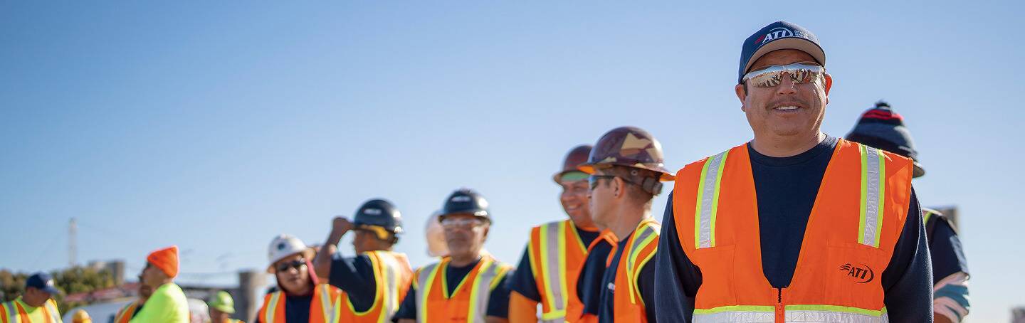 A smiling construction worker in an orange safety vest and hard hat, with other workers in the background.