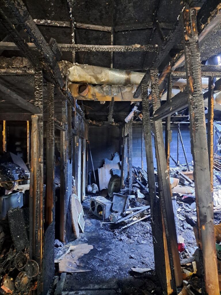 High-angle view of a fire-damaged interior, showing charred wood beams, debris, and a soot-covered floor.